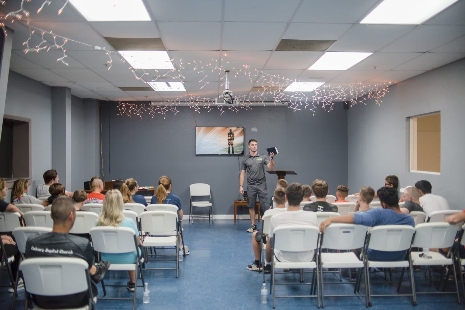 A speaker teaching from the front of the FUSION student room, with string lights overhead and students seated in rows