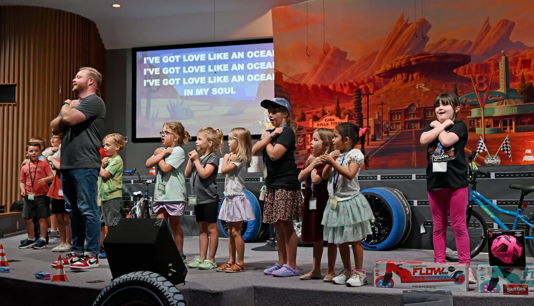 A Kids City leader on stage leading a room of children in singing, with the song words on the screens behind him
