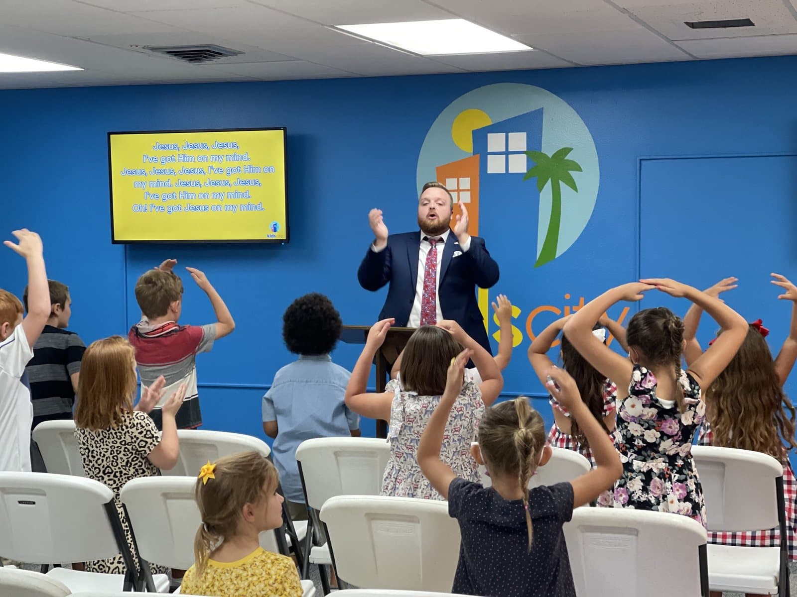 A leader in a shirt and tie leading young children in motions to a song in the Kids City room