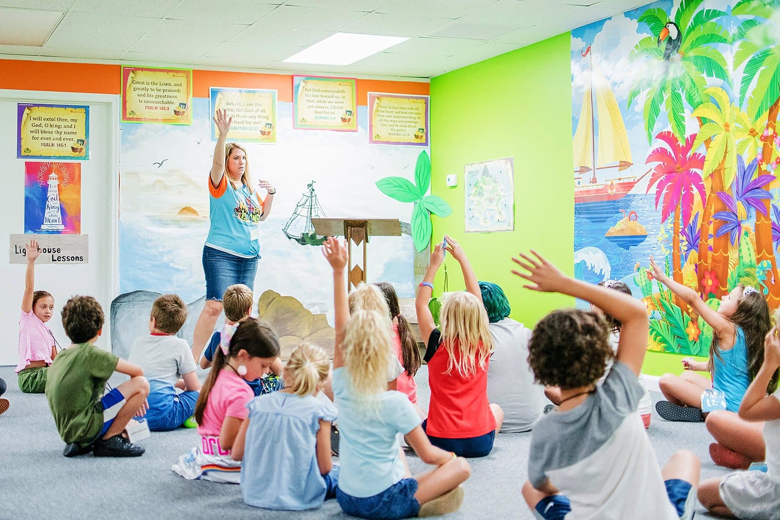 Children seated on the floor of a classroom with a painted palm-tree mural, reaching their hands toward their teacher