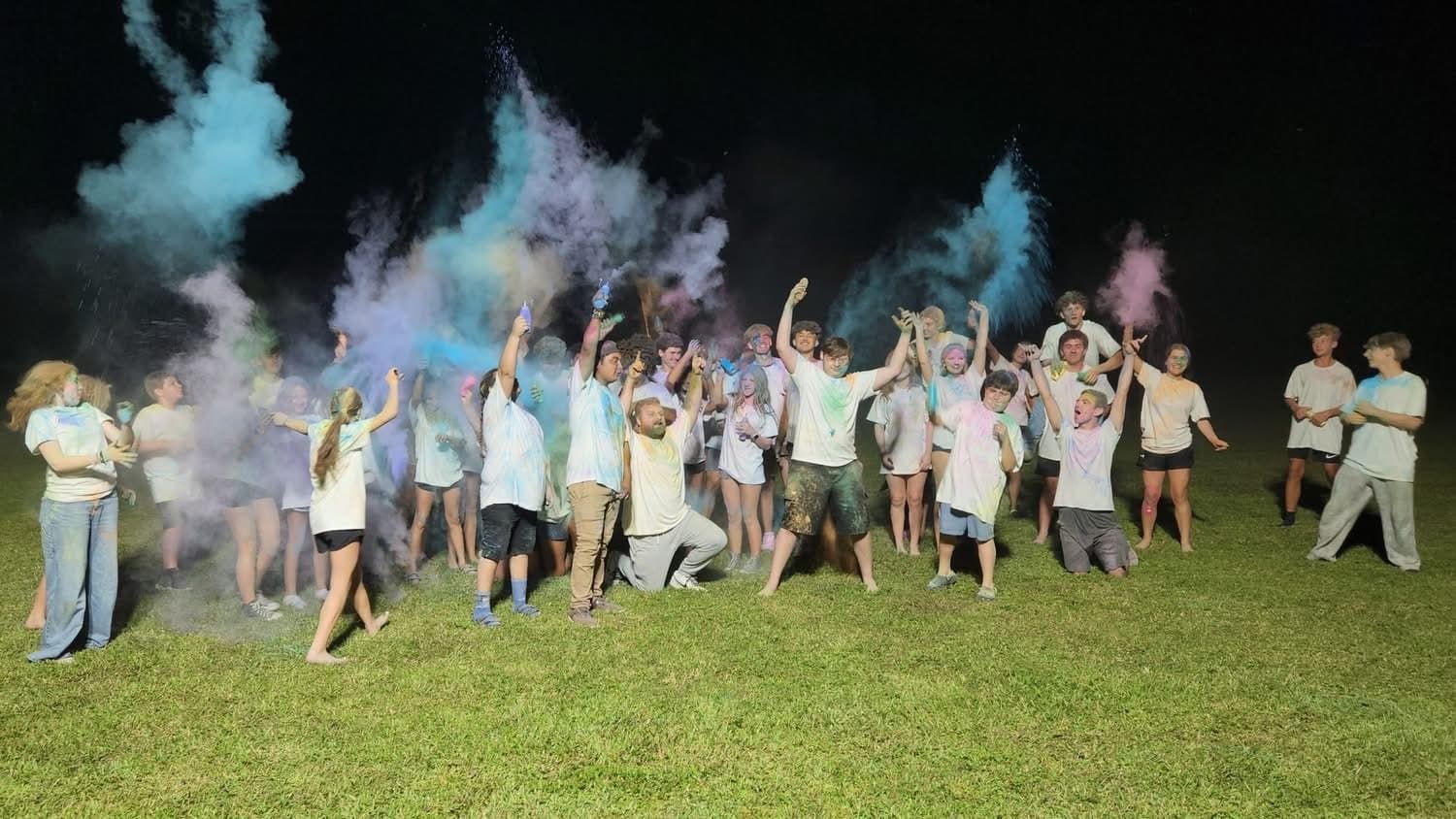 A line of teenagers in a grassy field throwing clouds of blue and pink colored powder into the air